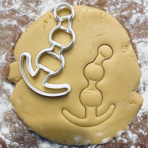 May include: A white plastic cookie cutter in the shape of a stylized anchor with a series of round balls on the top. The cookie cutter is on top of a round piece of cookie dough.