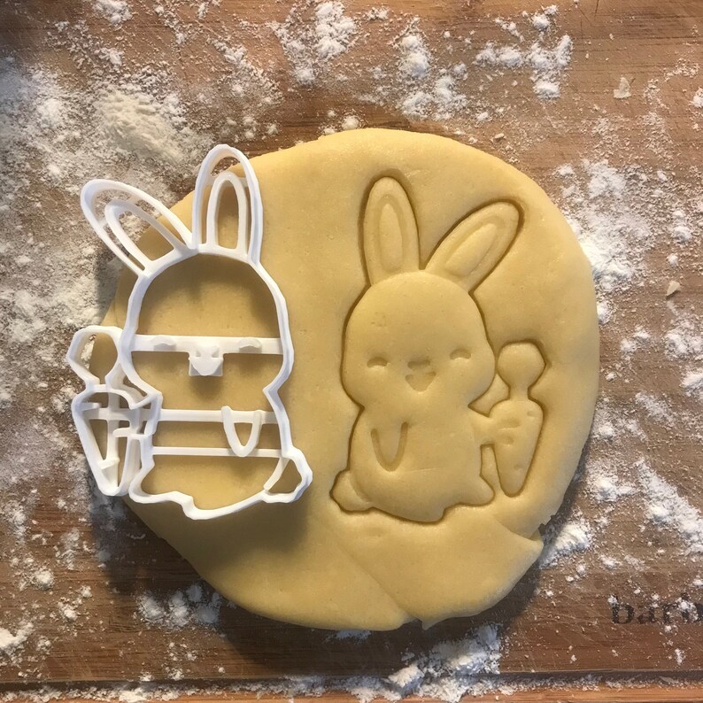 May include: A white plastic cookie cutter shaped like a bunny holding a carrot, next to a cookie dough cutout of a bunny holding a carrot.