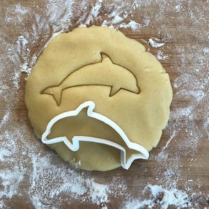 May include: A white plastic dolphin cookie cutter on top of a round piece of cookie dough. The cookie dough is light brown and has a dolphin shape cut out of it.