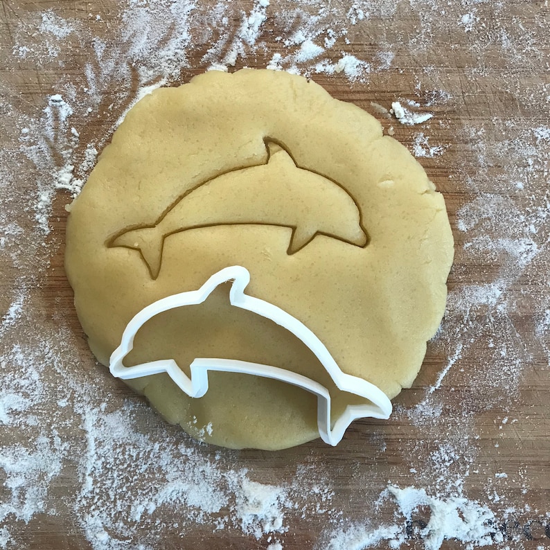 May include: A white plastic dolphin cookie cutter on top of a round piece of cookie dough. The cookie dough is light brown and has a dolphin shape cut out of it.