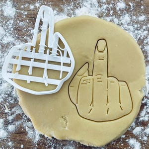 May include: A white plastic cookie cutter in the shape of a middle finger. The cookie cutter is on top of a round, light brown cookie dough.