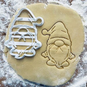Puede incluir: Un cortador de galletas de plástico blanco en forma de gnomo con un sombrero a rayas, y un recorte de masa de galleta marrón del mismo diseño de gnomo.