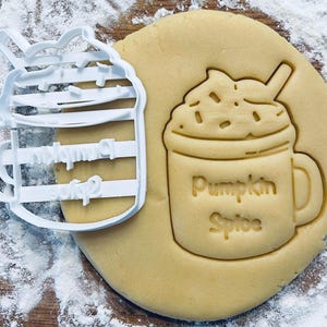May include: A white plastic cookie cutter shaped like a pumpkin spice latte with a straw and whipped cream. The cookie cutter is next to a cookie that has been cut out using the cookie cutter. The cookie is light brown and has the words "Pumpkin Spice" written on it.