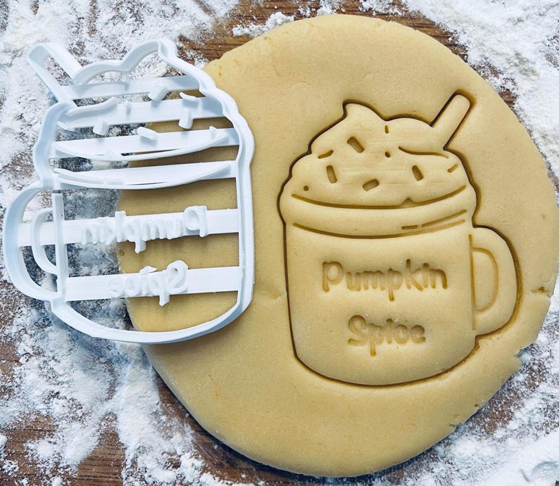 May include: A white plastic cookie cutter shaped like a pumpkin spice latte with a straw and whipped cream. The cookie cutter is next to a cookie that has been cut out using the cookie cutter. The cookie is light brown and has the words "Pumpkin Spice" written on it.