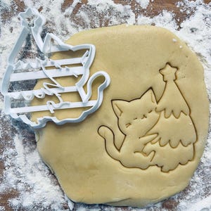 Può includere: Un tagliapasta di plastica bianca a forma di gatto che tiene un albero di Natale. Il tagliapasta è sopra un impasto per biscotti rotondo di colore marrone chiaro.