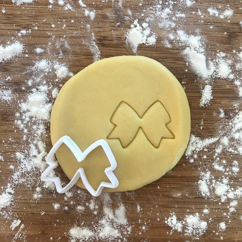 Pode incluir: Um cortador de biscoitos de pl&aacute;stico branco em forma de la&ccedil;o, em cima de uma pe&ccedil;a redonda de massa de biscoito n&atilde;o cozida.