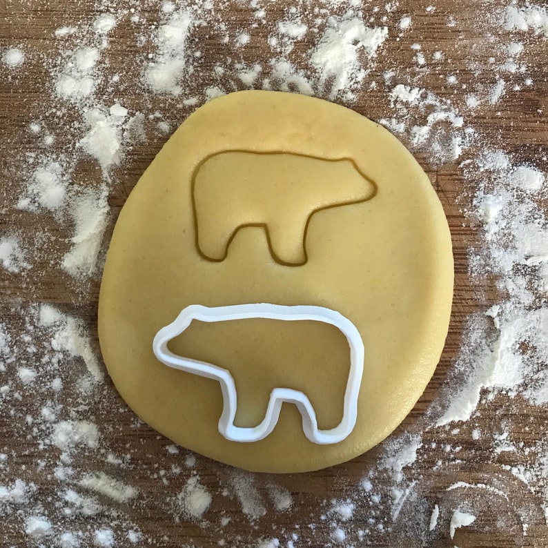 以下が含まれることがあります： 丸いクッキー生地の上に置かれたクマの形をしたクッキーカッターのクローズアップ。クッキーカッターは白で、生地は薄い茶色です。