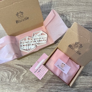 May include: A brown cardboard box with a pink tissue paper lining. The box contains two white plastic cookie cutters shaped like houses with a striped pattern. The box is labeled "Biscutie" and includes a thank you note.