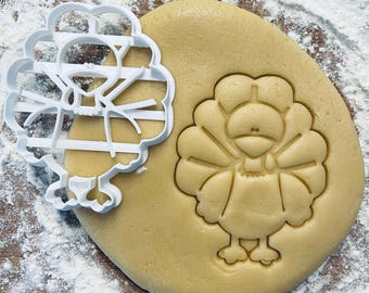 May include: A white plastic cookie cutter shaped like a turkey with a detailed design. The cutter is used to create a turkey-shaped cookie on a piece of dough.