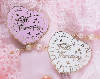 Puede incluir: Dos pines en forma de corazón con el texto "Frill Therapy" en letras doradas. Los pines están sobre un fondo de encaje rosa y blanco.