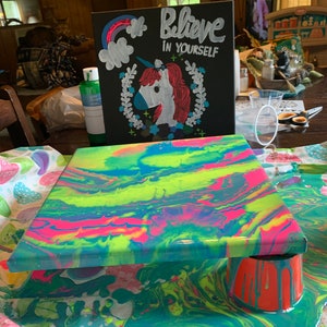 May include: A colorful abstract painting on a canvas with a bright green, blue, pink, and yellow color scheme. The painting is sitting on a table with a white tablecloth with a black sign that says "Believe in Yourself" with a unicorn illustration.