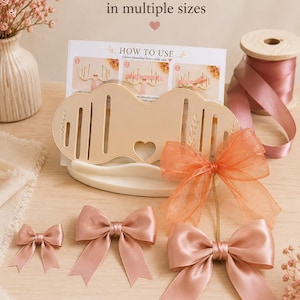 May include: A beige plastic bow-making tool with vertical slots and a central heart cutout, accompanied by an instructional card titled "HOW TO USE" and several finished satin and organza bows in shades of dusty pink and orange.