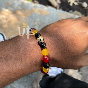 May include: A black, red, and yellow beaded bracelet with a gold soccer ball charm. The bracelet is made of round beads and has a simple design.
