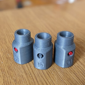 Op de afbeelding: Drie cilindrische grijze 3D-geprinte adapters met schroefdraad aan de bovenkant. Elk onderdeel heeft een kleine rode ronde inzet aan de zijkant en een vierkant logo in reliëf bij de basis. De onderdelen hebben een zichtbare gelaagde textuur.