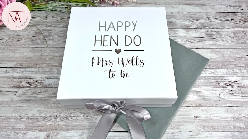 May include: A white gift box with the text "HAPPY HEN DO Mrs Wells to be" in black lettering. The box is tied with a silver ribbon and sits on a gray surface. The background is a light wood grain.