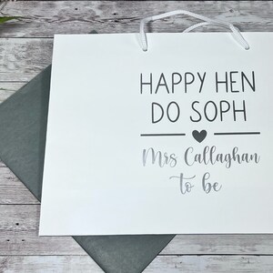 May include: White gift bag with the text "HAPPY HEN DO SOPH" and "Mrs Callaghan to be" in black and silver lettering. The bag has white handles and a heart symbol. A gray envelope is visible.