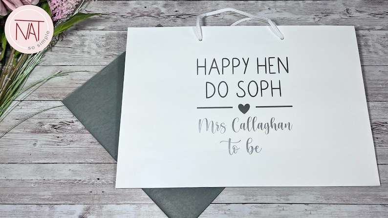 May include: White gift bag with the text "HAPPY HEN DO SOPH" and "Mrs Callaghan to be" in black and silver lettering. The bag has white handles and a heart symbol. A gray envelope is visible.