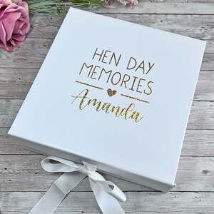 May include: White gift box with a ribbon closure. The box lid features gold glitter text that reads "HEN DAY MEMORIES Amanda" with a heart symbol. The box is on a wooden surface with a pink rose.