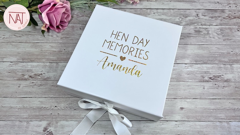 May include: White gift box with a ribbon closure. The box lid features gold glitter text that reads "HEN DAY MEMORIES Amanda" with a heart symbol. The box is on a wooden surface with a pink rose.