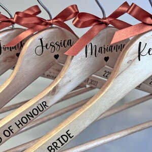 May include: Wooden hangers with names and titles in black script, each topped with a rust-colored ribbon bow. The hangers are personalized with names like "Jessica" and titles like "Bridesmaid". The date "22nd October 2022" is also printed.