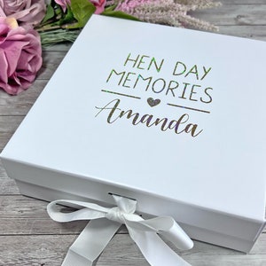May include: A white gift box with a lid, tied with a white ribbon. The box is decorated with the text "HEN DAY MEMORIES Amanda" in a metallic green font. The box is on a wooden surface.