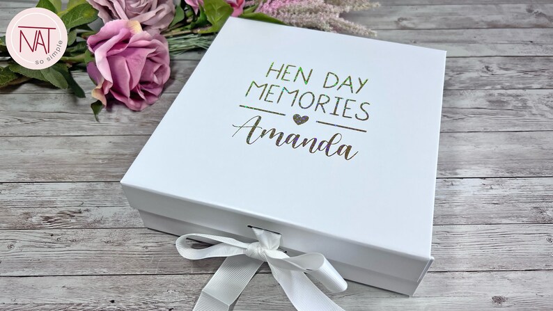 May include: A white gift box with a lid, tied with a white ribbon. The box is decorated with the text "HEN DAY MEMORIES Amanda" in a metallic green font. The box is on a wooden surface.