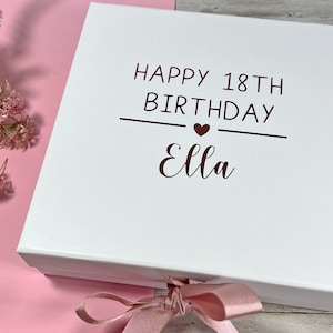 May include: A white gift box with the text "HAPPY 18TH BIRTHDAY Ella" in brown. The box is decorated with a pink ribbon and is next to pink and white flowers. The logo "NAT so simple" is in the upper left corner.