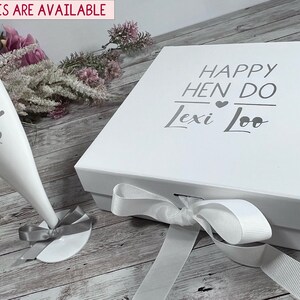 May include: A white gift box and champagne flute with the text "Happy Hen Do Lexi Loo" and "Lexi Loo" printed on them. The flute has a gray ribbon tied around the base. The box is tied with a white ribbon. Matching glasses are available.