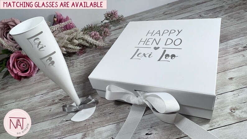 May include: A white gift box and champagne flute with the text "Happy Hen Do Lexi Loo" and "Lexi Loo" printed on them. The flute has a gray ribbon tied around the base. The box is tied with a white ribbon. Matching glasses are available.