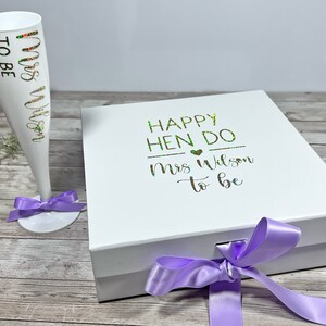 May include: A white champagne flute with "Mrs Wilson to be" in gold and green lettering, and a matching white gift box with "Happy Hen Do Mrs Wilson to be" text. Both items are decorated with purple ribbons, ideal for a bridal shower.