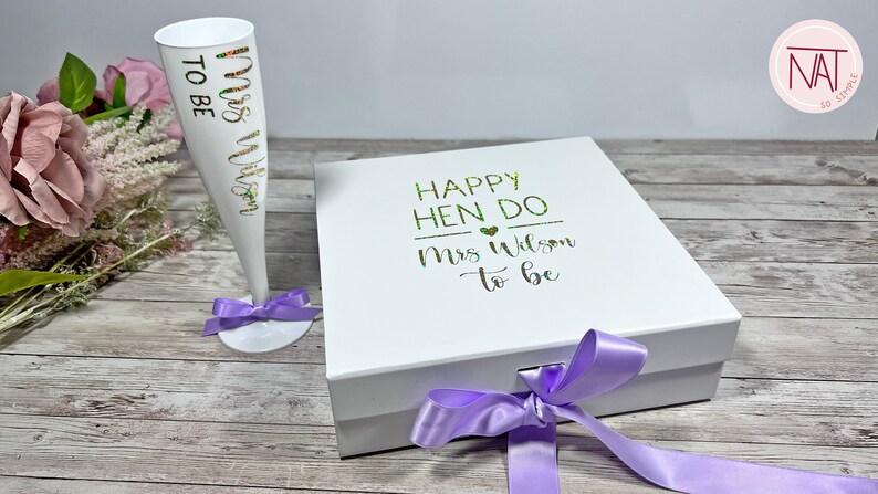 May include: A white champagne flute with "Mrs Wilson to be" in gold and green lettering, and a matching white gift box with "Happy Hen Do Mrs Wilson to be" text. Both items are decorated with purple ribbons, ideal for a bridal shower.