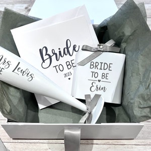 May include: A white gift box with a selection of bridal shower items. Includes a champagne flute, a small box, and a card, all with the words "Bride To Be" and "Mrs Lewis" printed on them. The items are arranged on gray tissue paper.