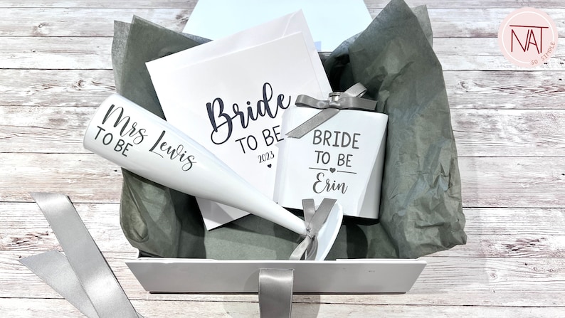 May include: A white gift box with a selection of bridal shower items. Includes a champagne flute, a small box, and a card, all with the words "Bride To Be" and "Mrs Lewis" printed on them. The items are arranged on gray tissue paper.