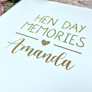 May include: White keepsake box with gold foil lettering that reads "HEN DAY MEMORIES Amanda". The text is in a script font with a heart symbol. The box is likely for a bachelorette party.