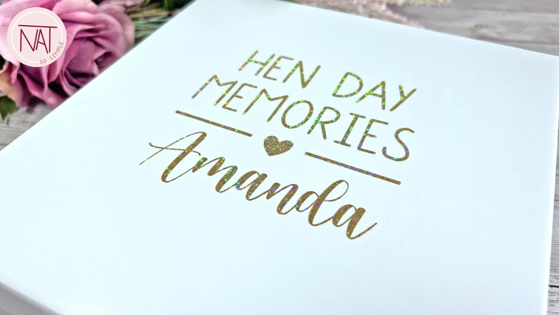 May include: White keepsake box with gold foil lettering that reads "HEN DAY MEMORIES Amanda". The text is in a script font with a heart symbol. The box is likely for a bachelorette party.