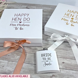 May include: Two white gift boxes with lids, tied with ribbons, and a flask. The boxes have the text "HAPPY HEN DO" and a name. The flask says "BRIDE TO BE" and a name. The background is a light wood surface.