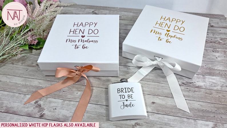 May include: Two white gift boxes with lids, tied with ribbons, and a flask. The boxes have the text "HAPPY HEN DO" and a name. The flask says "BRIDE TO BE" and a name. The background is a light wood surface.