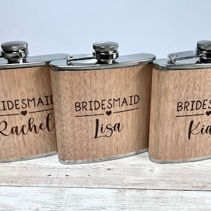 May include: Three wooden flasks with silver tops. Each flask is engraved with the word "Bridesmaid" above a name: Rachel, Lisa, and Kiah. The flasks have a light brown wood grain finish and are designed for a wedding party.