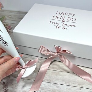 May include: A white gift box and champagne flute with rose gold text and accents. The box reads "Happy Hen Do Mrs. Traynor to be". The flute is inscribed with "Mrs. Traynor". A pink ribbon and bow are also present.