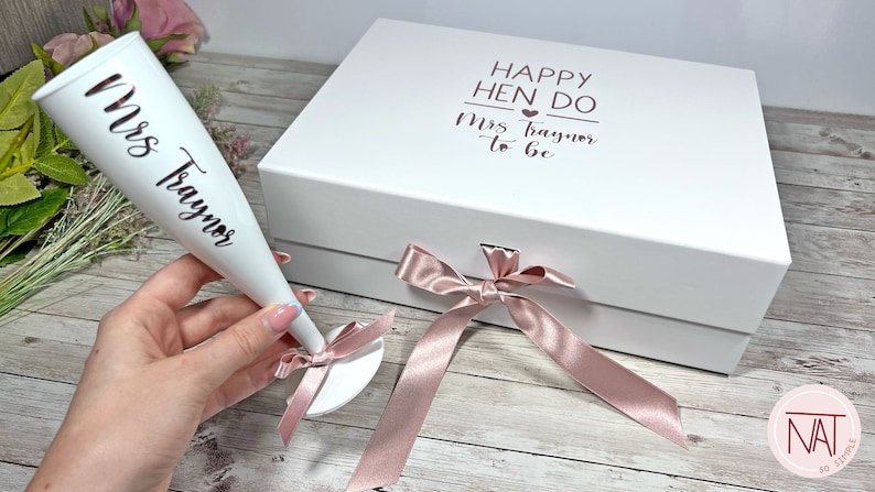 May include: A white gift box and champagne flute with rose gold text and accents. The box reads "Happy Hen Do Mrs. Traynor to be". The flute is inscribed with "Mrs. Traynor". A pink ribbon and bow are also present.