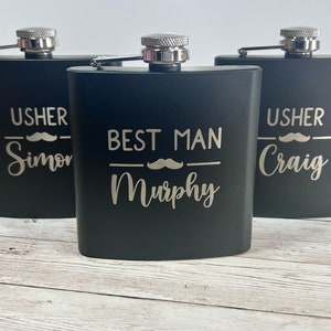 May include: Three black flasks with silver tops are displayed on a light wood surface. The flasks are engraved with the words "USHER Simon", "BEST MAN Murphy", and "USHER Craig", each with a mustache graphic.