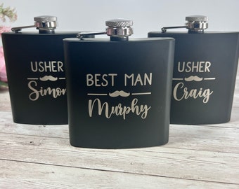 May include: Three black flasks with silver tops are displayed on a light wood surface. The flasks are engraved with the words "USHER Simon", "BEST MAN Murphy", and "USHER Craig", each with a mustache graphic.