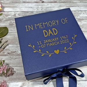 May include: A navy blue memory box with gold text reading "IN MEMORY OF DAD" and dates. The box is tied with a navy blue ribbon. Pink and white floral arrangements are in the background.