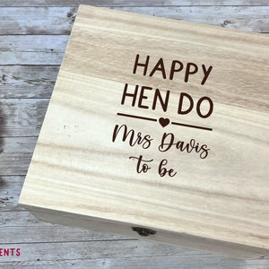 May include: A light-colored wooden box with the text "HAPPY HEN DO Mrs Davis to be" engraved on the lid. The box measures 24 inches wide, 24 inches long, and 10 inches high. A pink rose and other flowers are in the background.