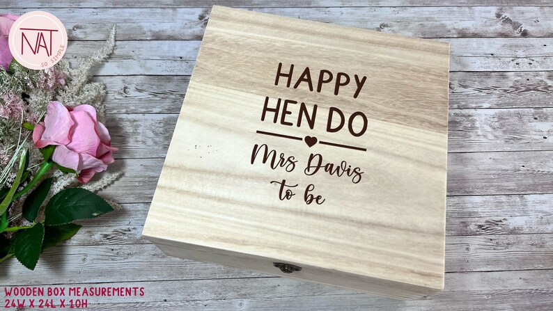 May include: A light-colored wooden box with the text "HAPPY HEN DO Mrs Davis to be" engraved on the lid. The box measures 24 inches wide, 24 inches long, and 10 inches high. A pink rose and other flowers are in the background.