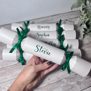 May include: White party crackers tied with green ribbons. Each cracker has a name printed on it in green script. The names visible are Nanny, Sophia, Milly, and Stevie. The crackers are on a wooden surface.