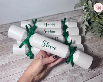 May include: White party crackers tied with green ribbons. Each cracker has a name printed on it in green script. The names visible are Nanny, Sophia, Milly, and Stevie. The crackers are on a wooden surface.