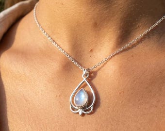 May include: A silver necklace featuring a teardrop-shaped moonstone pendant set within an open, stylized metal frame. The pendant hangs from a delicate silver chain, showcasing the iridescent blue glow of the polished gemstone.