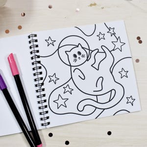 May include: A spiral-bound coloring book open to a page with a black and white illustration of a cat in space surrounded by stars. Two markers, one pink and one purple, are on the left. The background is a light wood surface with scattered confetti.
