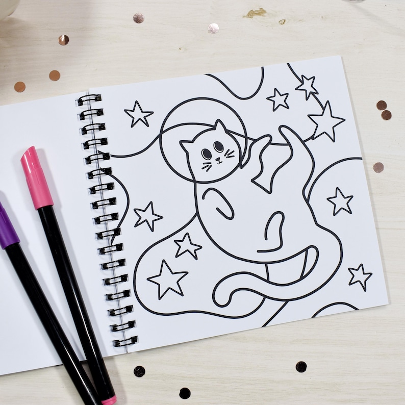 May include: A spiral-bound coloring book open to a page with a black and white illustration of a cat in space surrounded by stars. Two markers, one pink and one purple, are on the left. The background is a light wood surface with scattered confetti.